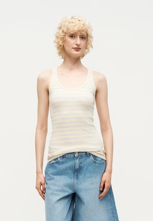 Jong persoon met krullend blond haar, die een licht gestreept tanktop en losse blauwe spijkerbroek draagt, staand tegen een effen achtergrond.