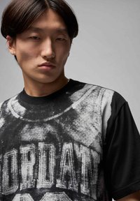 Czarny T-shirt z krótkim rękawem z szarą grafiką przedstawiającą wzór koszykówki, z wyraźnie widocznym słowem "JORDAN" na środku.