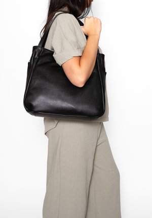 Donna in abito beige che porta una grande borsa tote in pelle nera sulla spalla, su sfondo bianco.