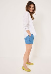 Camicia bianca a maniche lunghe, pantaloni corti in denim blu e ballerine gialle con un dettaglio a fiocco. Il modello è in piedi di lato, con le mani in tasca, su uno sfondo neutro.