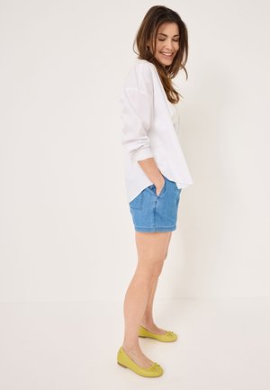 Chemise blanche à manches longues, short en denim bleu, et ballerines jaunes avec un détail en nœud. Le modèle se tient de profil, les mains dans les poches, sur un fond neutre.