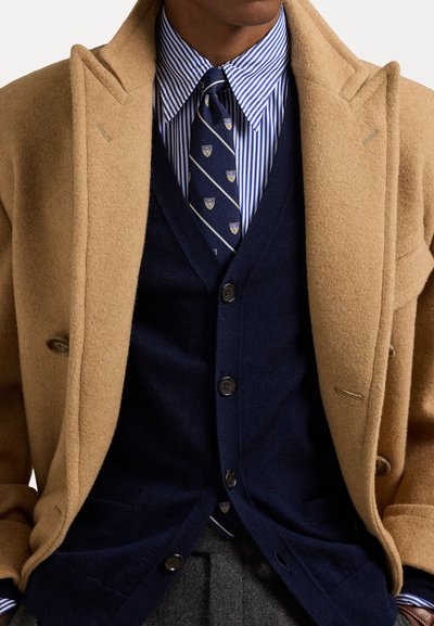 Camel wool blend overcoat, navy cardigan, striped blue shirt, patterned tie, detailed tailoring, buttons visible, layered textures.