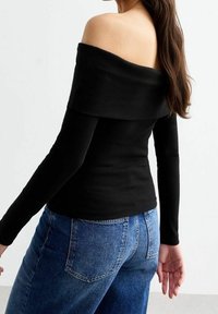 Top lungo a maniche lunghe nero off-the-shoulder con texture a coste, design aderente, abbinato a jeans in denim blu a vita alta.