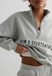 Gray sweatshirt with a high collar, zip closure, and bold black text "AIM'N SPORTSWEAR" on the front. The material appears soft and textured.