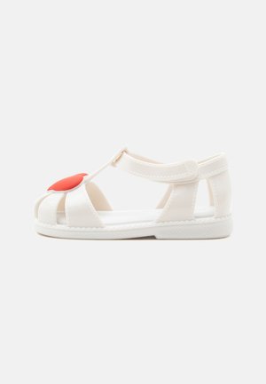 Sandale blanche pour tout-petit avec bout fermé, bride en T, fermeture à Velcro à la cheville et décoration en forme de cœur rouge à l'avant.
