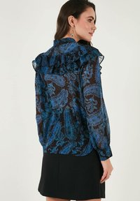 Blue paisley-patterned blouse made of sheer fabric with ruffled collar and long sleeves, paired with a black skirt.