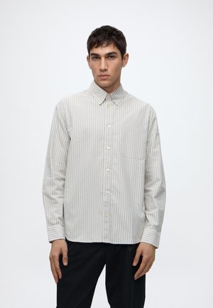 Joven con una camisa de rayas beige claro de manga larga con botones y pantalones negros, de pie frente a un fondo claro y simple.