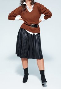 Femme portant un pull marron par-dessus une chemise blanche, une jupe plissée en cuir noire, une ceinture noire avec une boucle dorée et des bottines noires.