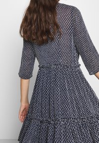 Marinblå klänning med ett lagerkjol, prydd med ett delikat vitt mönster. Långa ärmar och resår i midjan för en avslappnad passform.