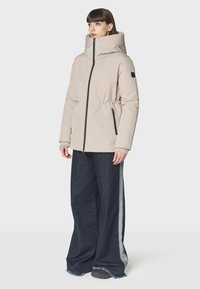 Chaqueta acolchada beige con cuello alto, cremallera frontal y cintura con cordón, combinada con unos vaqueros de pierna ancha de mezclilla oscura y zapatos negros.