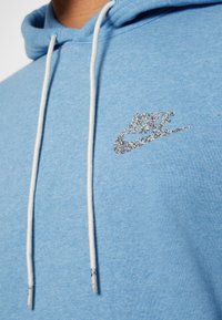 Ljusblå huvtröja med dragsnören, med en silverglittrande Nike-logotyp på bröstet och en mjuk textur.
