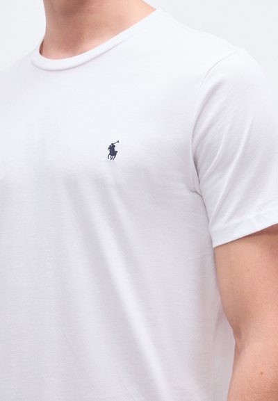 T-shirt branca de algodão com decote redondo, apresentando um pequeno logótipo bordado em azul-marinho no lado esquerdo do peito. Mangas curtas, textura suave.