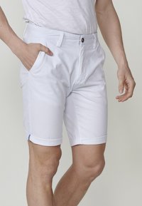 Homme portant un short blanc à ourlet retourné, une main dans sa poche, et un t-shirt blanc, sur fond uni.