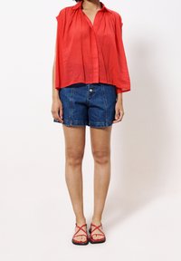 Persona con una blusa roja sin mangas de corte amplio, pantalones cortos de mezclilla azules y sandalias planas rojas con tiras, de pie contra un fondo blanco liso.