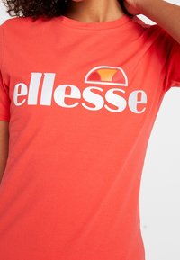 Korálové tričko s krátkým rukávem vyrobené z bavlny, se velkým bílým logem "ellesse" a barevným půlměsícovým akcentem nad ním.