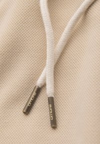 Gros plan sur un tissu beige avec deux cordons tressés blancs, chacun terminé par des embouts métalliques gravés "OMBRE."