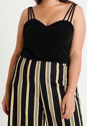 Mujer que lleva una camiseta negra sin mangas con triple tirante y pantalones negros de talle alto con rayas verticales amarillas y blancas.