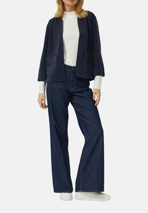 Frau trägt eine marineblaue Jacke mit offenem Schnitt über einem weißen Langarmshirt, dunkle Jeans mit weitem Bein und weiße Turnschuhe, steht vor einem schlichten Hintergrund.