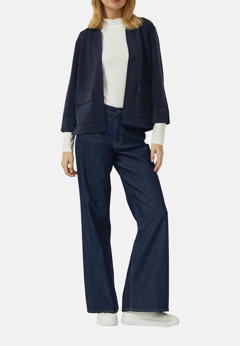 Žena na sebe má tmavomodrý otvorený kabátik cez biele tričko s dlhým rukávom, tmavé široké rifle a biele tenisky, stojí pred jednoduchým pozadím.