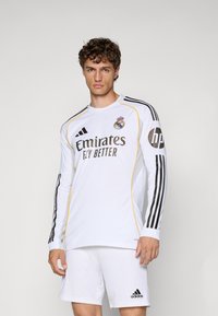 Jersey bianco a maniche lunghe con accenti neri e gialli, che presenta il logo "Emirates Fly Better", dettagli a strisce nere e branding Adidas.