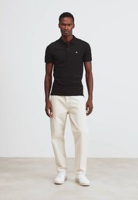 Camiseta polo negra de manga corta, con dos botones y un pequeño logo. Combinada con pantalones color crema y zapatillas blancas. Textura suave.