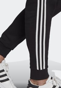Pantaloni sportivi neri con tre strisce verticali bianche lungo il lato, dotati di un polsino aderente. Abbinati a scarpe sportive bianche e nere.