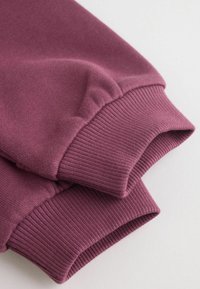Gros plan des manches pliées d'un sweat-shirt bordeaux avec des poignets côtelés sur un fond clair.