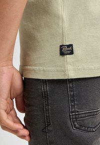Lichtgroene katoenen T-shirt met korte mouwen, voorzien van een blauw en gouden merklabel aan de zoom. In combinatie met donkergrijze denim jeans.