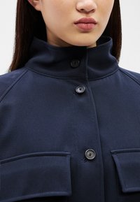 Tânără purtând o jachetă de culoare albastru închis, cu guler înalt, nasturi negri și două buzunare cu clapetă în față, văzută de la bărbie până la piept.