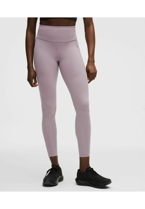 Kobieta w wysokich, liliowych legginsach przyciętych powyżej kostek i czarnych butach sportowych, stojąca na jednolitym jasnym tle.