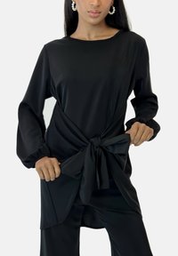Sort tunika i glat stof med lange ærmer, der har en knyttet detalje ved taljen og et afslappet fit design.