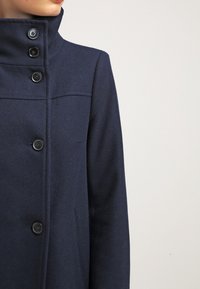 Cappotto in lana navy con colletto alto e quattro bottoni neri. Presenta un design sartoriale con una texture morbida e dettagli minimalisti.