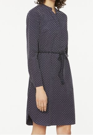 Vrouw draagt een marineblauwe jurk met lange mouwen en kleine witte patronen, gestrikt in de taille met een dunne riem.