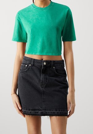 Femme portant un t-shirt court vert à manches courtes associé à une mini-jupe en jean noire taille haute avec un ourlet effiloché.