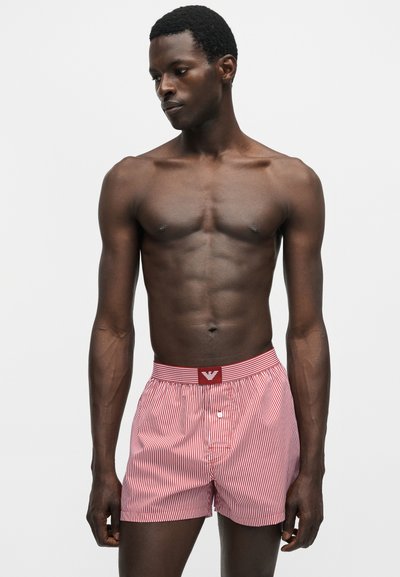 Ανδρικό μοντέλο με σκούρο δέρμα που φορά κόκκινα και λευκά ριγέ μποξεράκια σε απλό ανοιχτό γκρι φόντο.