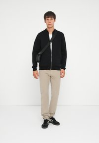 Veste bombardier noire zippée, t-shirt blanc, pantalon beige, baskets noires. Le mannequin tient un petit sac bandoulière noir, se tenant sur un fond blanc.