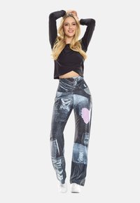 Blondīne sieviete melnā ilgrokāsas kultūraugā un zilās džinsās ar raibu stilu un rozā sirdi, pozējot ar paceltām rokām, smaida.