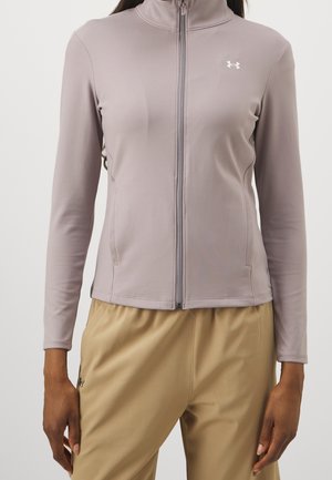 Femme portant une veste zippée violet clair avec un logo et un pantalon beige, debout devant un fond uni.