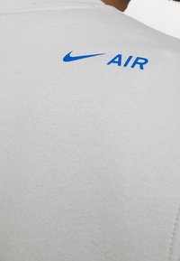 Ljusgrå t-shirt med en mjuk textur. Har en blå Nike-logga och texten "AIR" på den övre delen av baksidan. Närbild.