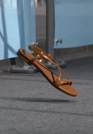 Καφέ δερμάτινο σανδάλι με λεπτούς ιμάντες και αγκράφα που αιωρείται πάνω από μια γκρι ασφάλτινη επιφάνεια κοντά σε ένα μπλε εμπόδιο ή φράχτη.