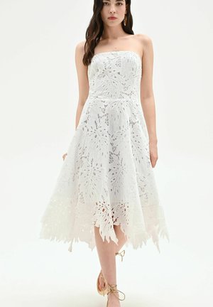 Femme portant une robe bustier blanche en dentelle avec des motifs de feuilles et un ourlet asymétrique, associée à des talons beiges à lacets, debout sur fond blanc.