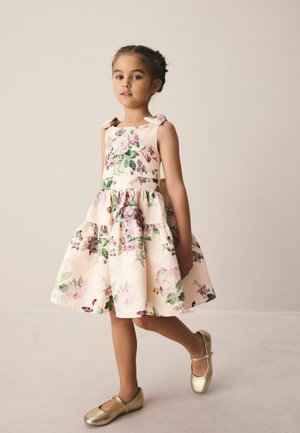 Junges Mädchen, das in einem cremefarbenen Blumenkleid mit Schleifendetails und goldenen Mary-Jane-Schuhen vor einem schlichten Hintergrund läuft.