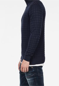 Mörkblå stickad tröja med ett strukturerat rutmönster, rundad halsringning och ribbad nederkant, i kombination med blå denimjeans.