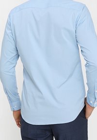 Camicia a maniche lunghe azzurro chiaro con una texture liscia, polsini abbottonati e orlo curvo, dal design pulito e semplice.