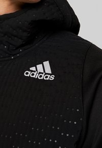 Sudadera con capucha negra con superficie texturizada, que presenta pequeños puntos. Logotipo de Adidas en blanco prominente en el pecho, con acentos de tela suave.