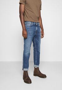 Vaqueros azules de mezclilla con un ajuste relajado, dobladillos remangados y un sutil detalle descolorido. Combinados con una camiseta ajustada marrón y botines marrones.