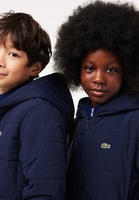 Des vestes rembourrées bleu marine avec capuches présentent une fermeture éclair à l'avant et le logo Lacoste, portées par deux enfants aux cheveux texturés, se tenant côte à côte.