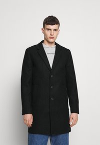 Abrigo negro texturizado con un diseño entallado, de un solo botón, que cuenta con tres botones y dos bolsillos frontales sobre una sudadera gráfica gris.