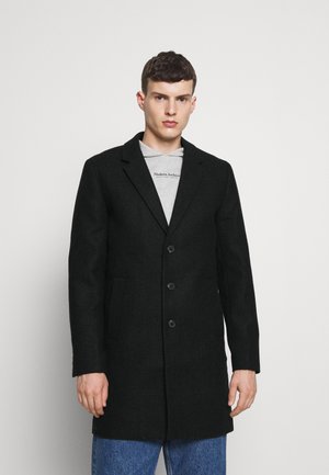 Abrigo negro texturizado con un diseño entallado, de un solo botón, que cuenta con tres botones y dos bolsillos frontales sobre una sudadera gráfica gris.