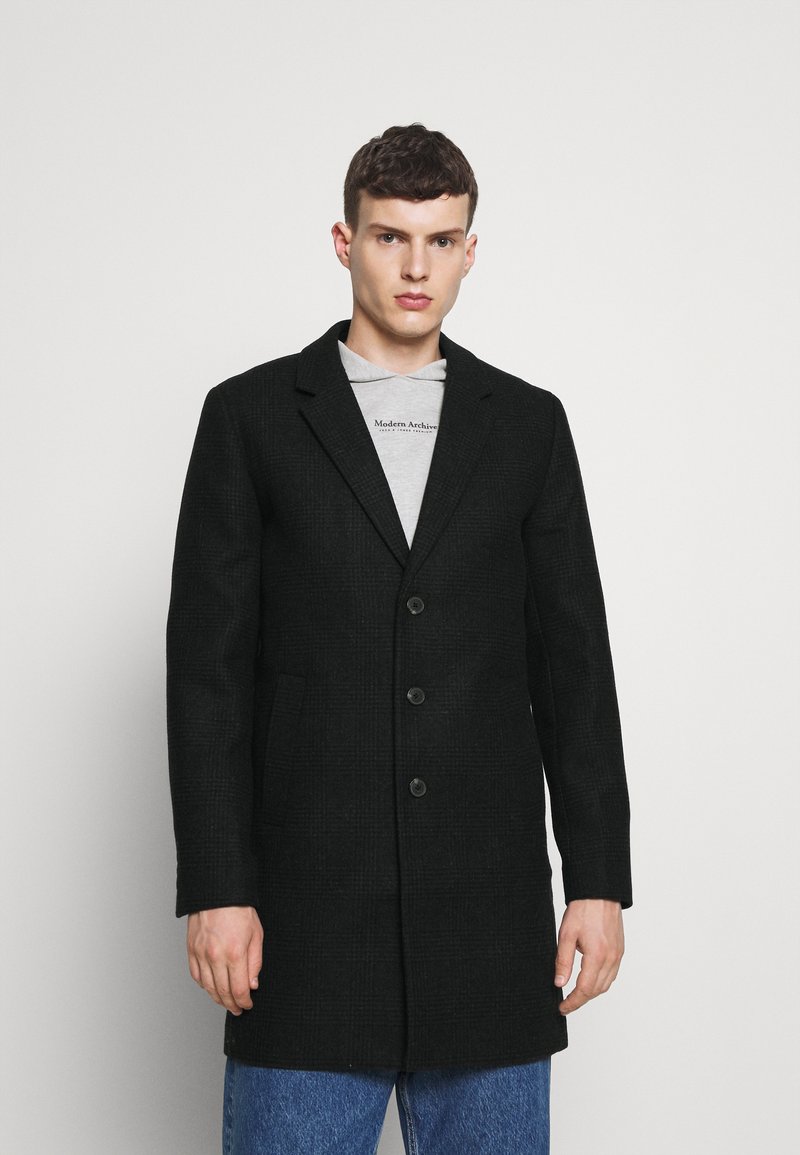 Abrigo negro texturizado con un diseño entallado, de un solo botón, que cuenta con tres botones y dos bolsillos frontales sobre una sudadera gráfica gris.